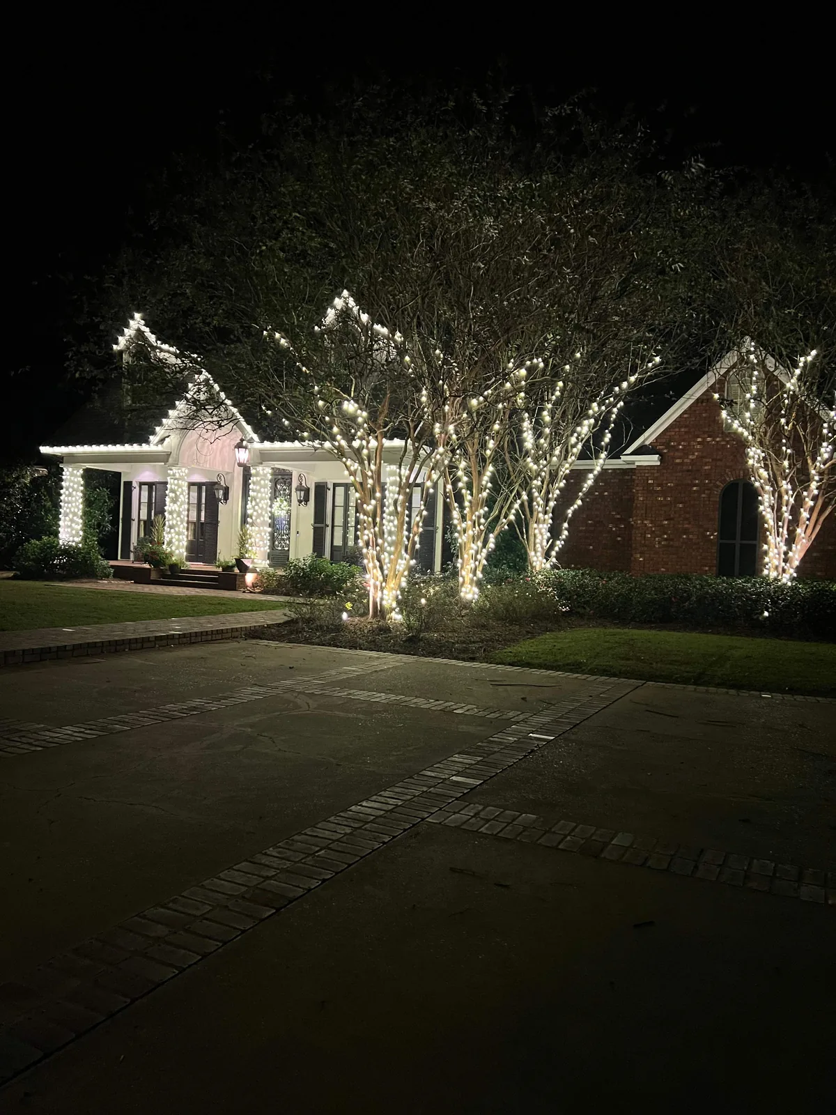 Christmas lights along a home’s roofline and porch columns, with illuminated trees beside the front walk at night
