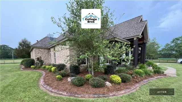 Curved landscape bed with brick curbing, layered plantings, boulders, and an ornamental tree in front of a stone home